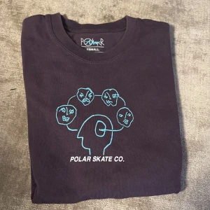 Svart t-shirt från Polar Skate Co. - Säljer en svart t-shirt från Polar Skate Co. med ett unikt tryck av ansikten i blått på framsidan. Perfekt för dig som gillar streetwear och vill ha något som sticker ut. T-shirten är kortärmad och gjord i mjukt material.