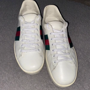 Gucci Ace skor  - Snygga vita sneakers från Gucci med klassiska gröna och röda ränder på sidan. Skorna har en rund tå och snörning framtill. Baktill finns en grön detalj med krokodilmönster. Perfekta för en stilren look.