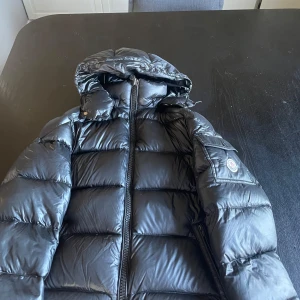 Svart dunjacka från Moncler - Säljer en svart dunjacka från Moncler med huva och dragkedja. Jackan har ett quiltat mönster och en snygg glansig finish. Perfekt för kalla dagar när du vill hålla dig varm och stilren.nfc funkar den kommer i en Moncler case.