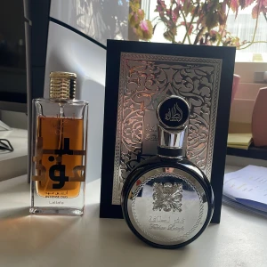 Fakjr Lattafa, Amer Al oud  - Två eleganta parfymflaskor från Lattafa. Den ena är en rektangulär flaska med bärnstensfärgad vätska och guldfärgad kork, märkt 'Intense Oud'. Den andra är en rund flaska med silverdetaljer och en dekorativ förpackning med orientaliskt mönster. Perfekt för den som älskar lyxiga dofter.
