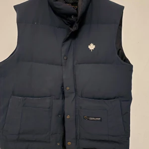 Mörkblå dunväst från Canada Goose - Säljer en mörkblå dunväst från Canada Goose med knappar och en broderad logotyp på bröstet. Västen har två stora fickor framtill och är perfekt för kyligare väder.