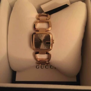 Guldig fyrkantig klocka från Gucci - Snygg klocka från Gucci med fyrkantig urtavla i guldton och stilren länk i metall. Urtavlan är silverfärgad med minimalistiska markeringar och datumruta. Länken har unika, öppna länkar som ger en modern vibe. Perfekt accessoar för dig som gillar lyxig och trendig stil.