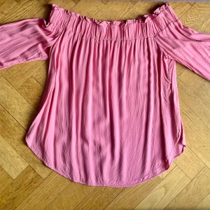 Rosa offshoulder blus med knytärmar - Supersöt rosa offshoulder blus med långa ärmar och knyt vid ärmsluten. Blusen har en rynkad, elastisk kant upptill och är tillverkad i ett lätt, luftigt tyg som faller snyggt. Perfekt för dig som gillar en romantisk och trendig stil.
