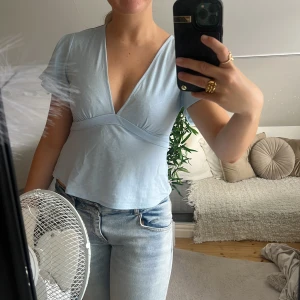 Ljusblå omlottopp med v-ringning rosett i bak - Supersöt ljusblå omlottopp med djup v-ringning och korta ärmar. Toppen har en markerad midja och är tillverkad i mjuk bomull, perfekt för en chill och fräsch look. Passar snyggt till jeans eller kjol.