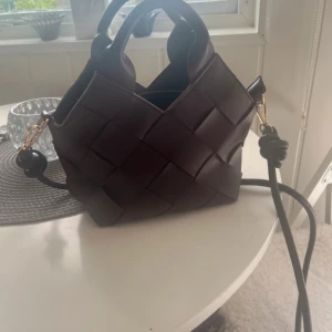Pitaya väska - Säljer en snygg brun axelväska i flätad design med korta handtag och justerbar axelrem. Väskan är gjord i skinn och har guldfärgade metalldetaljer. Perfekt för dig som vill ha en trendig och stilren accessoar.