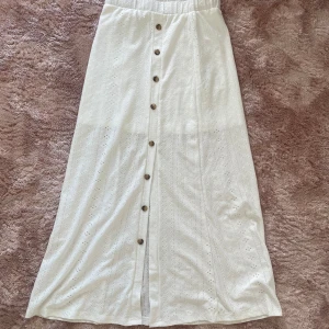 Vit långkjol med broderade detaljer - Superfin vit långkjol med broderade mönster och knappar framtill. Kjolen har elastisk midja och är fodrad för extra komfort. Perfekt för en somrig look med söt vintagekänsla och snygga detaljer längs hela kjolen.