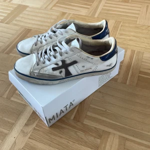 Vita sneakers från Premiata - Snygga vita sneakers från Premiata med blå detalj på hälen och svart logga på sidan. Klassisk låg modell med coola detaljer och tryck på sulan. Storleken är 44 och allt OG finns med. Vid minsta lilla fundering skriv gärna!