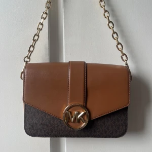 Brun axelväska från Michael Kors - Snygg axelväska från Michael Kors i brunt skinn och mörkbrunt mönstrat material. Väska är nästan som nytt skick, har endast tappat lite färg på inre knappen men de syns knappt i verkligenheten speciellt när väskan är stängd. Får plats med väldigt mycket:) 