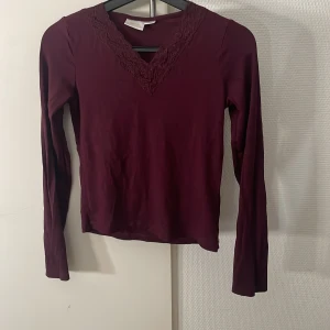 Vinröd långärmad topp från Gina Tricot - Vinröd långärmad topp från Gina Tricot med v-ringning och spetsdetaljer runt halsen. Snygg och enkel modell i mjukt material, perfekt att styla med jeans eller kjol. Passar dig som gillar en feminin touch på vardagslooken.