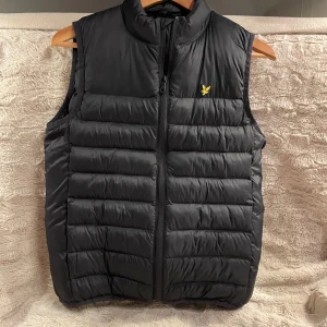 Svart dunväst från Lyle & Scott - Snygg svart dunväst från Lyle & Scott med gul broderad logga på bröstet. Västen har quiltad design, hög krage och dragkedja framtill. Perfekt att slänga över en hoodie för extra värme. Klassisk och stilren look.