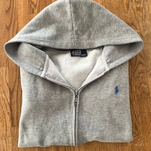 Grå hoodie från Polo Ralph Lauren - Säljer en grå hoodie från Polo Ralph Lauren med dragkedja och blå logga på bröstet. Tröjan har huva, långa ärmar och är tillverkad i mjuk bomullsmix. Perfekt för chill dagar eller när du vill ha en clean och enkel stil.
