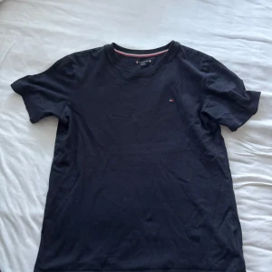 Svart t-shirt från Tommy Hilfiger - Klassisk svart t-shirt från Tommy Hilfiger med liten broderad logga på bröstet. T-shirten har rund halsringning med snygg randig detalj på insidan av kragen. Tillverkad i mjuk bomull och har korta ärmar. Perfekt för en clean och stilren look.