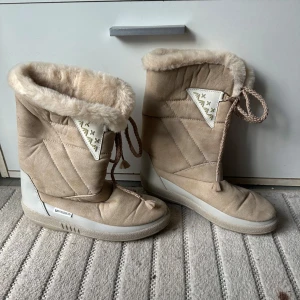 Beige moon boots med fuskpäls - Beiga moon boots från Top Line med mjuk fuskpäls runt skaftet och quiltade detaljer. Skorna har snörning framtill och en platt, räfflad sula. Perfekta för kalla vinterdagar och riktigt mysiga att ha på sig.