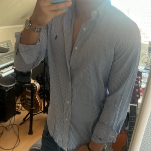 Randig skjorta från Ralph Lauren - Säljer en klassisk blåvit randig skjorta från Ralph Lauren med broderad logga på bröstet. Skjortan har lång ärm, knappar hela vägen och en snygg krage. Perfekt för dig som gillar stilrena och tidlösa plagg.