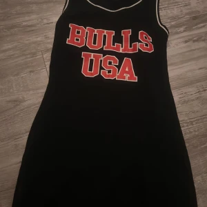 Svart sportig klänning Bulls USA Lemon - Svart kortklänning från Lemon med sportig känsla och vita kontrastkanter. Framsidan har stort rött tryck med texten 'BULLS USA'. Ärmlös modell, perfekt för en cool och avslappnad stil.