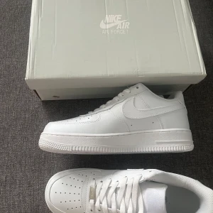 Nike Air Force 1 helvita sneakers - Klassiska Nike Air Force 1 i helvitt skinn med låg profil och perforerad tå. Snygg och tidlös modell med vit sula och diskret Nike-logga på sidan. Perfekta för dig som vill ha en clean och stilren look till vardags.