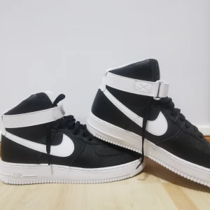 Nike Air Force 1'07 High svart/vit  - Helt ny/aldrig använts. Svarta och vita Nike Air Force 1 High sneakers med klassisk Swoosh-logga i vitt, högt skaft och justerbar rem runt ankeln. Skorna har snörning, platt sula och är tillverkade i präglat skinn för en clean och sportig look.