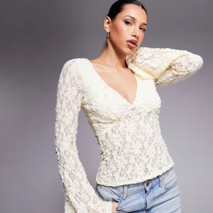 Gul spetsblus med v-ringning - Helt ny, oanvänd från ASOS. Romantisk vit blus i spets med djup v-ringning och långa, lätt utsvängda ärmar. Figurnära passform och transparent spetsmönster ger en feminin vibe. Perfekt att styla med jeans eller kjol för en trendig look.