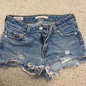 Levi's ljusblå jeansshorts low waist - Säljer ett par ljusblå jeansshorts från Levi's med låg midja och rak passform. Shortsen har slitna detaljer, fransig rå kant och klassiska fem fickor. Perfekta för sommaren och ger en avslappnad vibe.