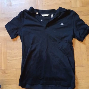 Svart pikétröja från Gant - Klassisk svart pikétröja från Gant med korta ärmar och liten broderad logga på bröstet. Tröjan har krage och knäppning med två knappar. Perfekt för en clean och stilren look. Hör av er vid närmaste fundering eller snabb affär. Pris kan diskuteras 