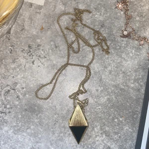 Guldigt halsband med svart diamantformad hänge - Unikt halsband med kedja i guldfärg och ett stort diamantformat hänge. Hänget har en cool kombination av guld och svart, där den svarta delen är blank och kontrasterar snyggt mot det borstade guldet. Perfekt statement-piece för dig som gillar edgy accessoarer. Ingen aning vart den är ifrån men är helt ny tror att den är rostfritt stål. OBS den är väldigt lång så går till brösten på mig som är 162 🫶🏻🫶🏻