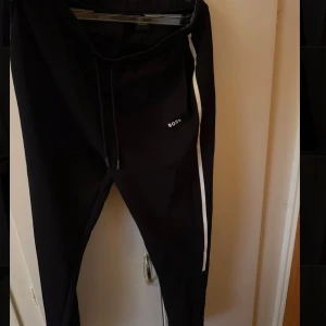 Svarta mjukisbyxor från Boss - Svarta mjukisbyxor från Boss med vit rand längs benet och diskret logga på framsidan. Byxorna har snörning i midjan och en bakficka. Perfekta för en chill och sportig stil.