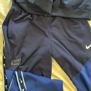Nike blå och svarta blåa  mjukisbyxor XL - Snygga mjukisbyxor från Nike i blått och svart med vita loggor längs sidorna. Byxorna har elastisk midja, sidofickor och är tillverkade i Dri-Fit polyester som andas. Perfekta för träning eller chill. Klassisk Nike-stil med sportig känsla.
