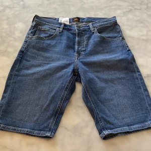 Blå jeansshorts från Lee - Snygga blå jeansshorts från Lee med fem fickor, gylf med dragkedja och knapp, samt snygga kontrastsömmar. Modellen är rak och slutar strax ovanför knäna. Helt nya med lappen kvar. Perfekta för en chill sommarstil. ☀️