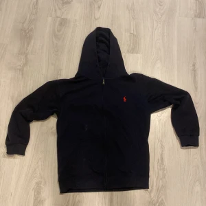 Svart hoodie från Polo Ralph Lauren - Klassisk svart hoodie från Polo Ralph Lauren med röd broderad logga på bröstet. Tröjan har dragkedja framtill, huva och två fickor. Tillverkad i mjuk bomull och har långa ärmar med muddar. Perfekt för en chill och stilren look.