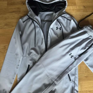 Grå vindjacka från Under Armour - Snygg grå vindjacka från Under Armour med svart logga på bröstet. Jackan har huva med snörning, svarta dragkedjor och ventilerande detaljer vid fickorna. Perfekt för träning eller chill dagar. Materialet är mjukt och lätt, och jackan har en sportig vibe.