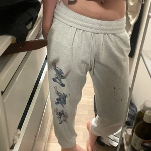 Grå Stitch mjukisbyxor från Disney - Grå mjukisbyxor från Disney med Stitch-tryck på ena benet. Byxorna har resår i midjan och muddar vid bensluten. Perfekta för chill dagar hemma och har en avslappnad passform. Den har lite hårfärg på låret men det är inget speciellt man tänker på.