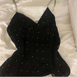 Fest topp - Supersnygg svart linnetopp från MNG Limited Edition med glittriga stenar och tunna kedjeaxelband. Toppen har en lätt draperad halsringning och är perfekt för dig som vill sticka ut. Passar till allt från jeans till kjol för en cool look.