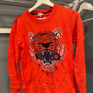 Röd Kenzo tröja med tigertryck - Cool orange tröja från Kenzo med ikoniskt broderat tigerhuvud och texten 'Kenzo Paris' framtill. Tröjan har rund halsringning och långa ärmar. Perfekt statement-piece för dig som vill sticka ut.