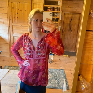 Röd blommig blus med spegeldekor - Säljer en transparent röd blus med blommigt mönster och inslag av rosa och orange. Blusen har v-ringning med dekorativa speglar och broderier framtill samt långa, vida ärmar. Perfekt för dig som gillar boho-vibe och färg.