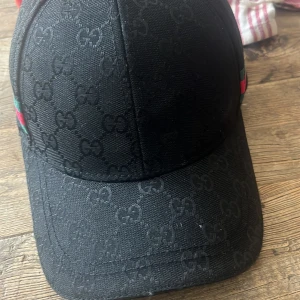 Svart keps från Gucci med GG-mönster - Snygg svart keps från Gucci med klassiskt GG-monogram över hela kepsen. Kepsen har böjd skärm och diskreta gröna och röda detaljer på sidorna. Tillverkad i bomull och polyester för en lyxig känsla och perfekt passform.