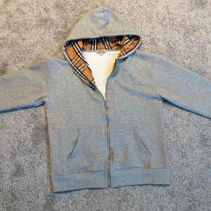  Burberry hoodie  - Snygg grå hoodie från Burberry med dragkedja och klassiskt rutigt foder i huvan. Jackan har broderad logga på bröstet, fickor fram och snörning i huvan. Perfekt för dig som gillar streetstyle och vill ha något som sticker ut. Pris kan såklart diskuteras