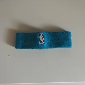 Turkos NBA pannband i frotté - Turkost pannband från NBA i mjuk frotté med broderad NBA-logga framtill. Perfekt för dig som gillar basket eller vill ha en sportig accessoar. Elastiskt material som sitter skönt runt huvudet.
