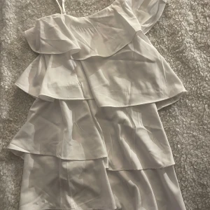 Vit volangklänning från Gina Tricot - Supersöt vit kortklänning från Gina Tricot med flera lager volanger och en axelbandslös design. Perfekt för sommarens alla tillfällen, lätt och luftig med snygg omlottkänsla. Stilren och enkel att matcha med accessoarer.