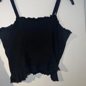 Svart smockad linnetopp med volang - Supersöt svart linnetopp med smockad struktur och justerbara axelband med knapp. Toppen har volangkant nertill och längs urringningen för en extra gullig touch. Perfekt till jeans eller kjol när du vill ha en chill men ändå snygg look. Går att justera banden eller ta av helt. ❤️passar S-xl 