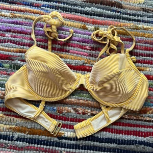 Gulvit randig bikiniöverdel med snörning - Superfin bikiniöverdel i gulvitt randigt mönster med vadderade kupor och bygel. Knytband i nacken och justerbart spänne i ryggen. Perfekt för stranden eller poolen, ger en fräsch och somrig look. Materialet känns mjukt och följsamt.