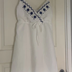Vit kortklänning med blå broderier🤍 - Söt vit kortklänning med blå broderade blommor. V-ringad framtill och smock där bak för bekväm pass form. Liten fläck där bak med men inget man tänker på och går säkert lätt bort💕 Nästintill helt oanvänd utöver provning!!🙌🏼