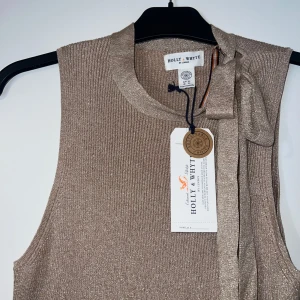 Glittrig beige ärmlös topp Holly & Whyte - Supersnygg glittrig beige ärmlös topp från Holly & Whyte. Ribbstickad med en cool knyt-detalj vid axeln som ger extra stilpoäng. Perfekt att styla med jeans eller kjol för en trendig look. Passar olika storlekar. Aldrig använd. ❤️