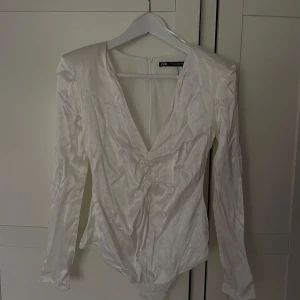 Vit body i satin från Zara - Vit body från Zara i glansigt satinliknande material. Djup v-ringning med snygg rynkdetalj och knyt framtill. Långärmad och med dragkedja i ryggen.
