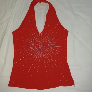 Röd halterneck topp med hjärtan - Röd halterneck topp med djup v-ringning och glittrande stenar i ett strålande mönster från mitten med hjärtan. Toppens material är stretchigt och passar perfekt för att sticka ut.