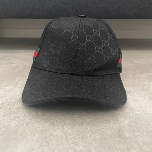 Svart keps från Gucci med monogram - Helt ny svart keps från Gucci med diskret GG-monogram över hela ytan. Klassisk böjd skärm och justerbar rem baktill. Sidorna har gröna och röda ränder som ger den ikoniska Gucci-looken. Tillverkad i bomull och polyester för en lyxig känsla.
