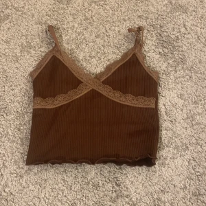 Brun ribbad linnetopp med spets H&M - Supersöt brun ribbad linnetopp från H&M Divided med tunna axelband och spetsdetaljer i en ljusare brun nyans. Croppad modell med vågig nederkant och omlott-effekt framtill. Perfekt för lager-på-lager eller till jeans.