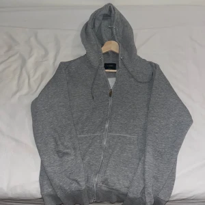 Grå hoodie från Jack & Jones - Mysig grå hoodie från Jack & Jones med dragkedja och huva. Tröjan har långa ärmar, snörning i huvan och två stora fickor framtill. Insidan är fodrad med mjukt material som gör den extra skön att ha på sig.