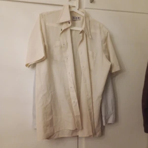 Beige kortärmad skjorta från N3P - Snygg beige skjorta från H3P med korta ärmar och klassisk krage. Skjortan har knappar framtill och bröstficka, perfekt för en avslappnad stil. Lätt och luftig, passar bra till varmare dagar.