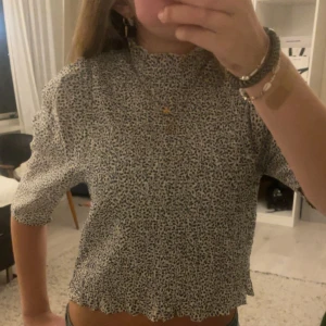 Blommig blus från H&M, vit/blå - Supersöt blommig blus från H&M i vitt och mörkblått. Modellen är croppad med vågig kant nertill och har trekvartsärm. Tunt och luftigt material, perfekt för vår och sommar. Blusen har hög hals och ett tätt blommönster 