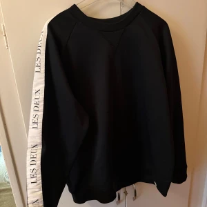 Svart sweatshirt Les Deux med vita detaljer - Svart sweatshirt från Les Deux med vita band längs ärmarna där det står 'LES DEUX' i svart text. Tröjan har rund halsringning och långa ärmar. Perfekt för en clean och sportig look.